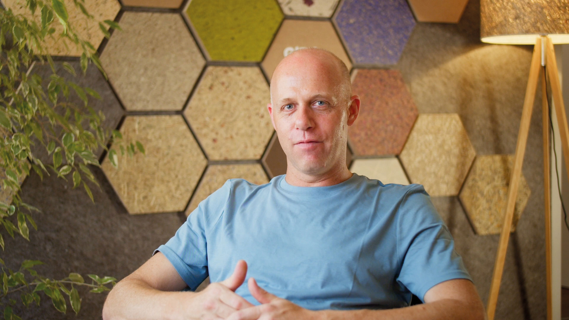 Mann in blauer T-Shirt sitzt vor einer Wand mit bunten Hexagon-Designs, neben einer Pflanze.