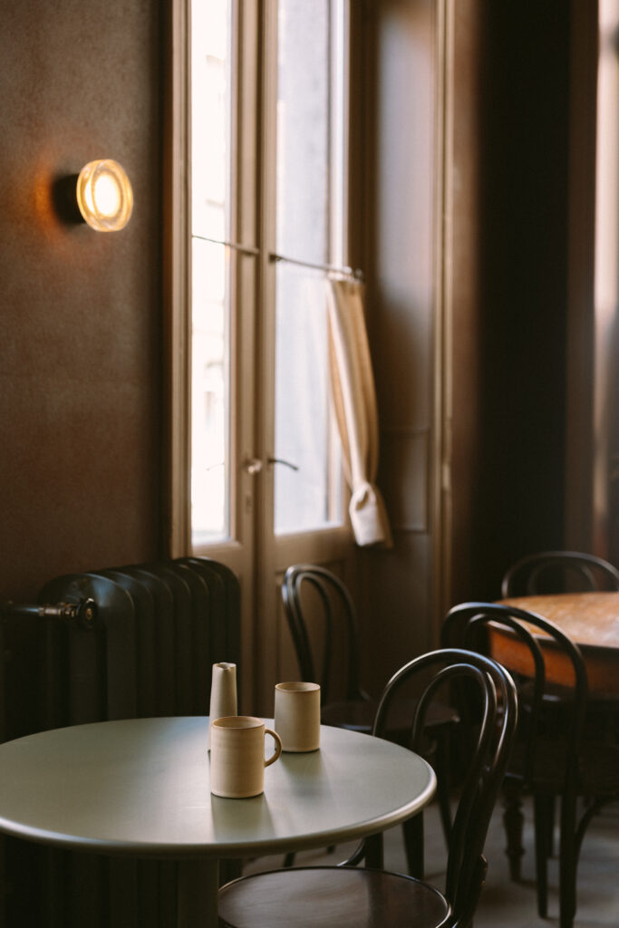 Ein heller Raum mit einem runden Tisch, drei Keramikbechern und einem Fenster mit Vorhängen