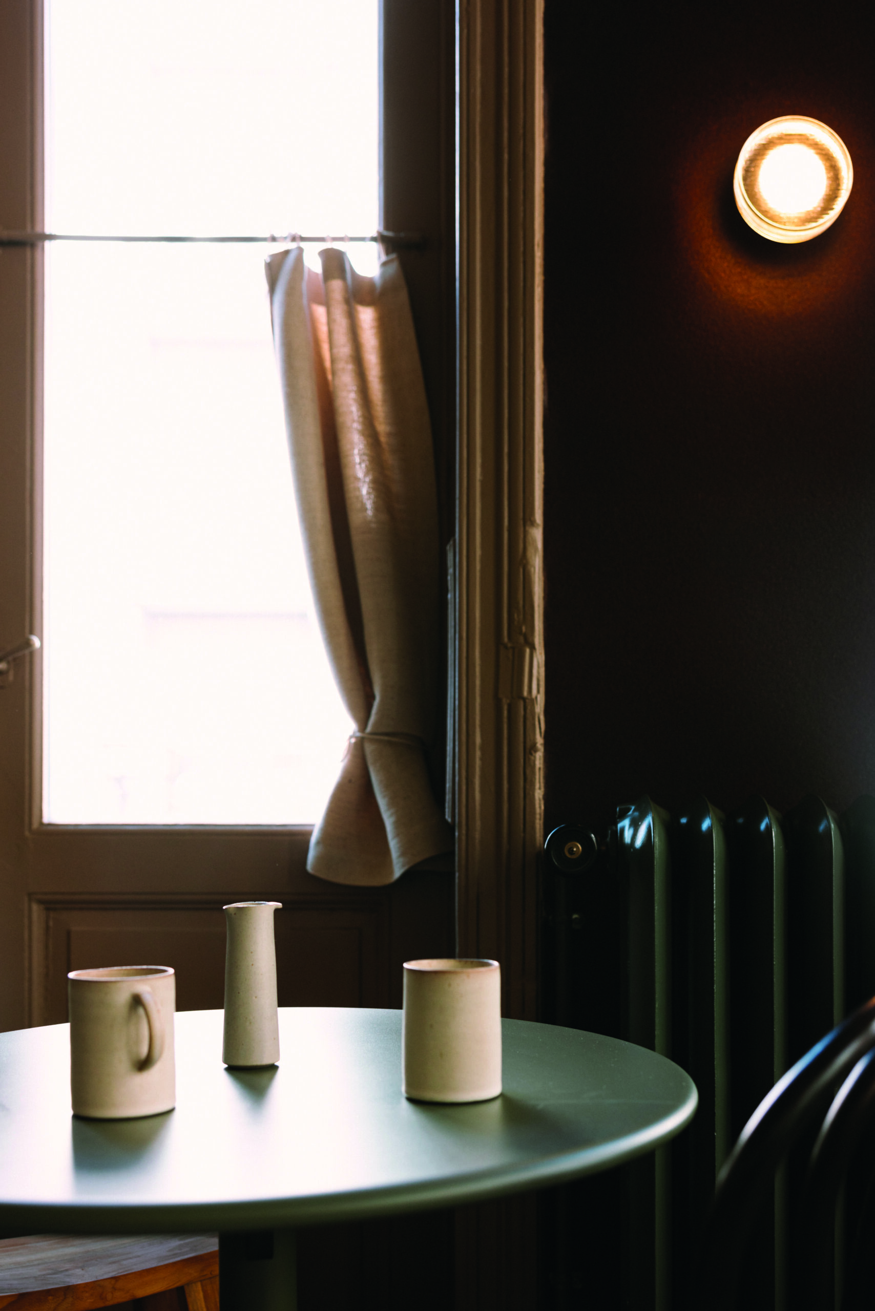 Fenster mit beige Vorhängen, beleuchtete Wandlampe, runde Tische mit weißen Keramikvasen.