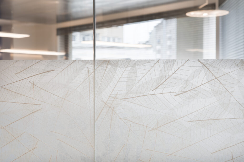 A glass wall with a leaf pattern below a modern office interior with large windows and ceiling ...