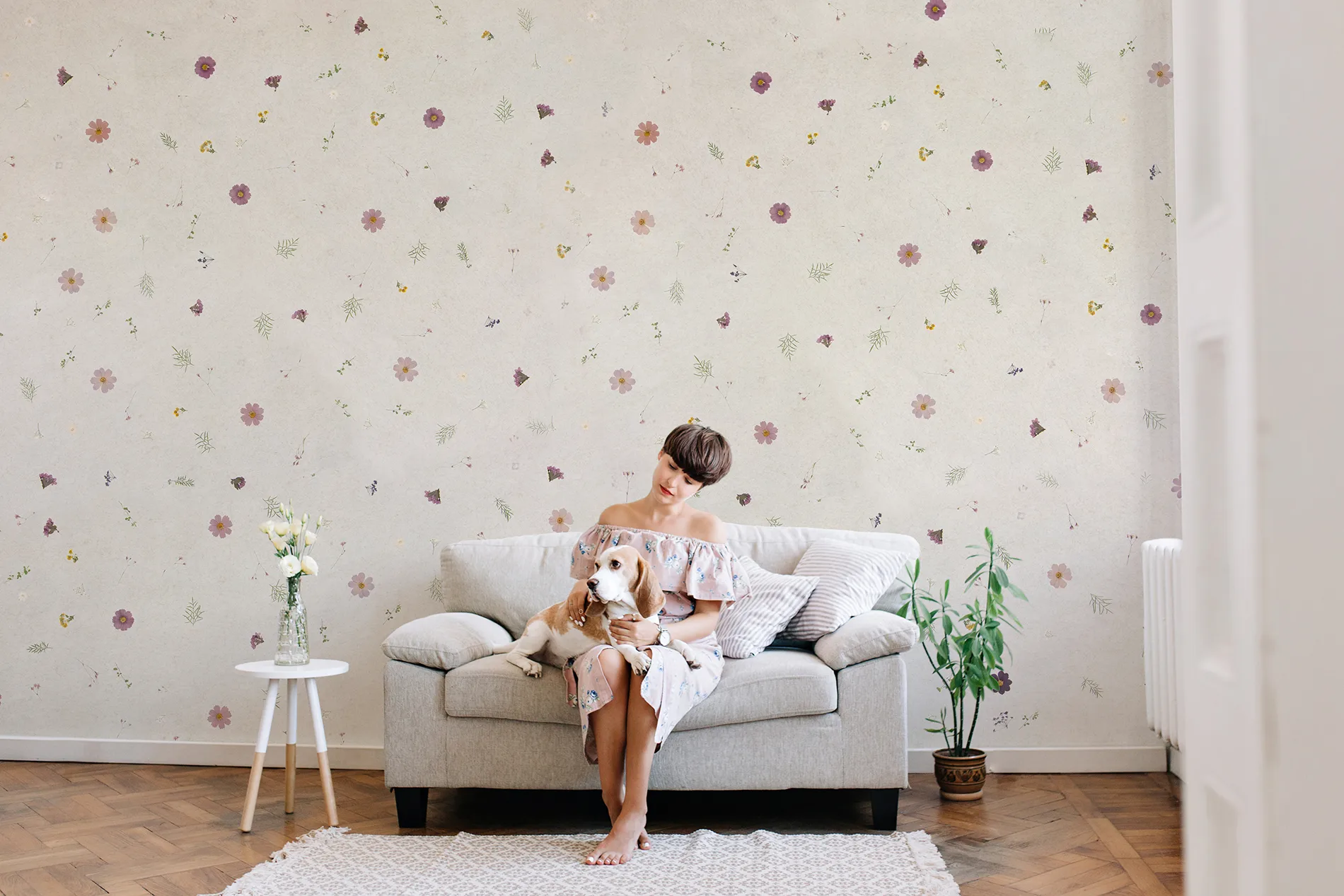 Frau sitzt auf Sofa mit Hund, im Raum mit Blumenmuster-Wand aus echten Blumen und Naturmaterialien und Pflanzen