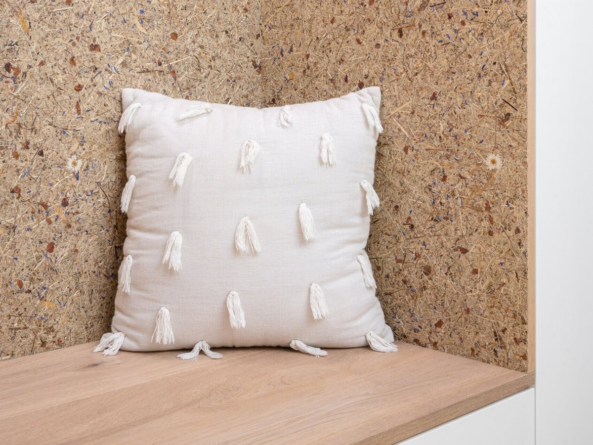 Weißes Kissen mit Fransen vor einer Holzbank und einer Wand aus gepresstem Holz.