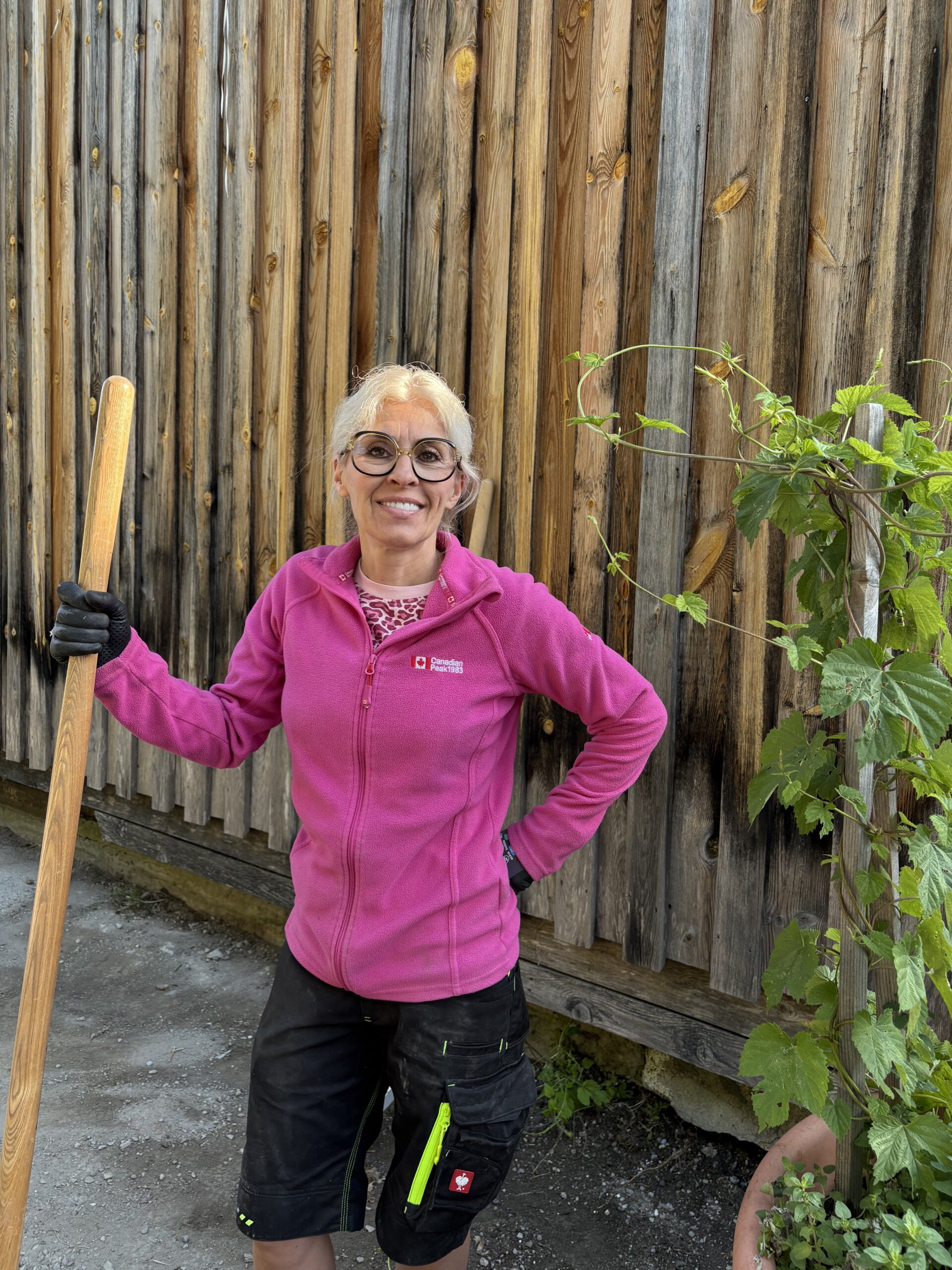 Eine Frau mit Brille und pinkem Pullover hält einen Gartenrechen vor einer Holzwand, umgeben vo...